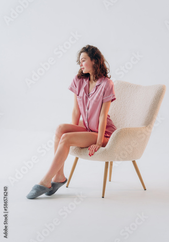 Beautiful young woman sitting in armchair on white background. Curly girl in pink silk pajama