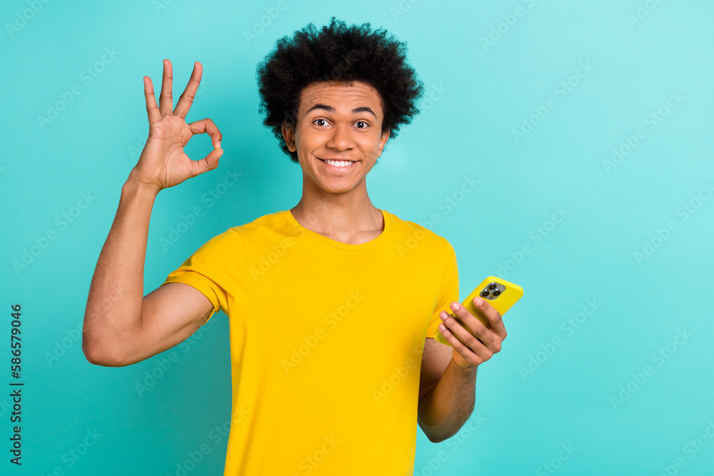 Portrait of optimistic satisfied guy wear yellow t-shirt showing okey holding smartphone isolated on vibrant teal color background Portrait of optimistic satisfied guy wear yellow t-shirt showing okey holding smartphone isolated on vibrant teal color background