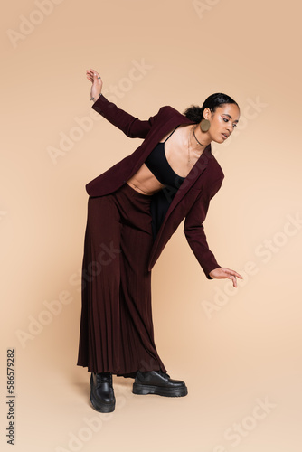 full length of charming african american woman in stylish maroon suit with wide leg pants posing on beige.