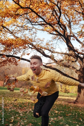 Happy guy throwing fall leaves in autumn park, joyful outdoors, arms outstretched. Funny man in yellow clothes having fun in nature. Concept of relax joy and leisure activity. Copy ad text space