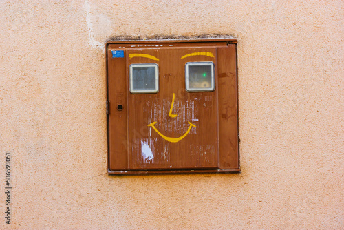 Industrial electrical junction box on the orange wall with a funny image in a form of a smiling face. Decorating objects and details of exterior of a building. Heat, light meter outdoor on a street.