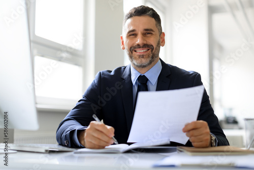 Happy middle aged businessman in suit working with documents at workplace in office and smiling at camera, free space © Prostock-studio