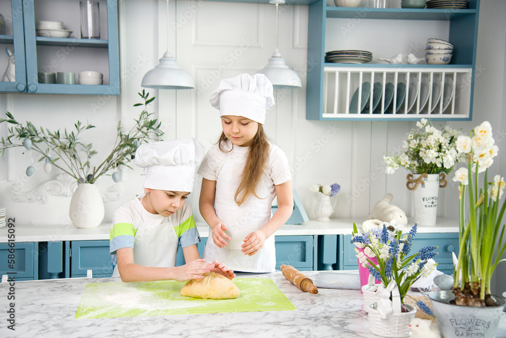 little cute funny smiling girl and boy wearing a white chef's hats and uniform kneading the dough for easter bread it in the blue and white kitchen with easter decorations little cute funny smiling girl and boy wearing a white chef's hats and uniform kneading the dough for easter bread it in the blue and white kitchen with easter decorations