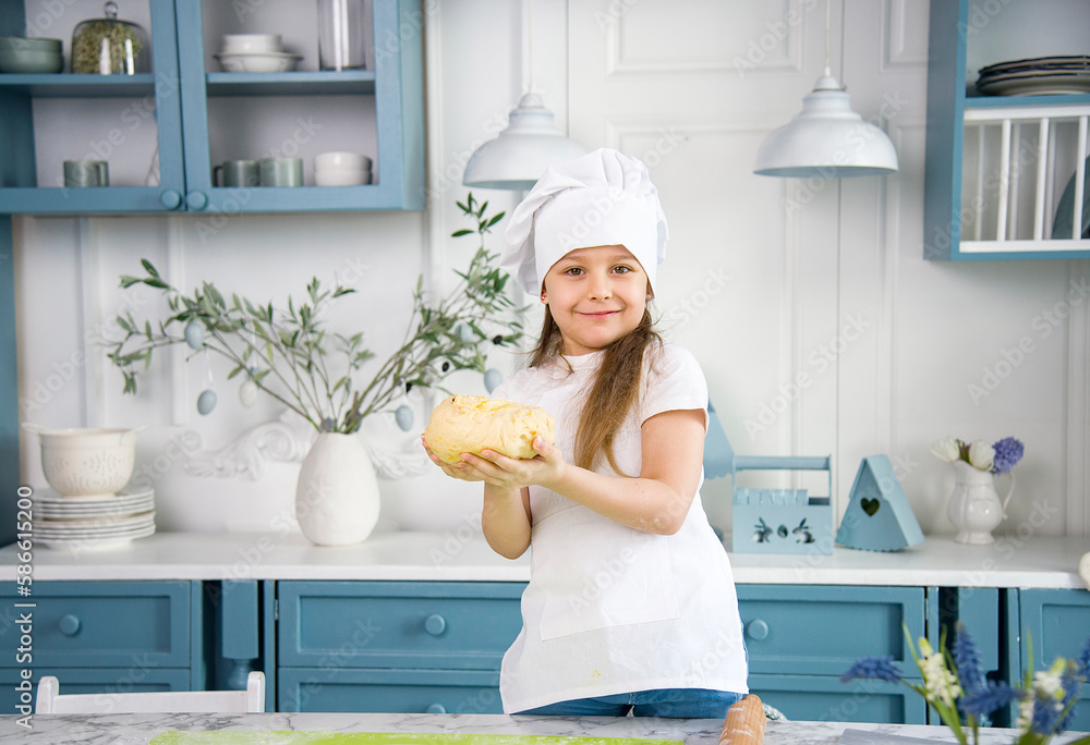 little cute smiling girl wearing a white chef's hat and uniform with tongue kneading the dough for cake in the blue and white kitchen with easter decoration little cute smiling girl wearing a white chef's hat and uniform with tongue kneading the dough for cake in the blue and white kitchen with easter decoration