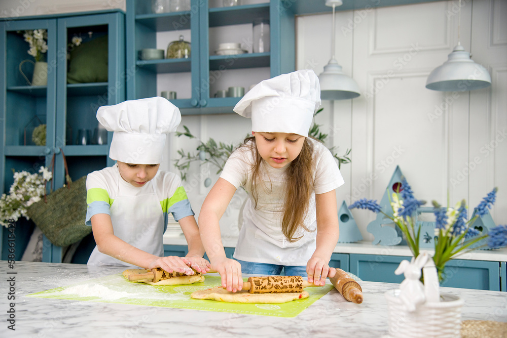 little cute funny smiling girl and boy wearing a white chef's hats and uniform kneading the dough for easter bread it in the blue and white kitchen with easter decorations little cute funny smiling girl and boy wearing a white chef's hats and uniform kneading the dough for easter bread it in the blue and white kitchen with easter decorations