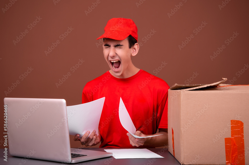 Funny courier in a red uniform sits at his desk. Brown background. Funny courier in a red uniform sits at his desk. Brown background.