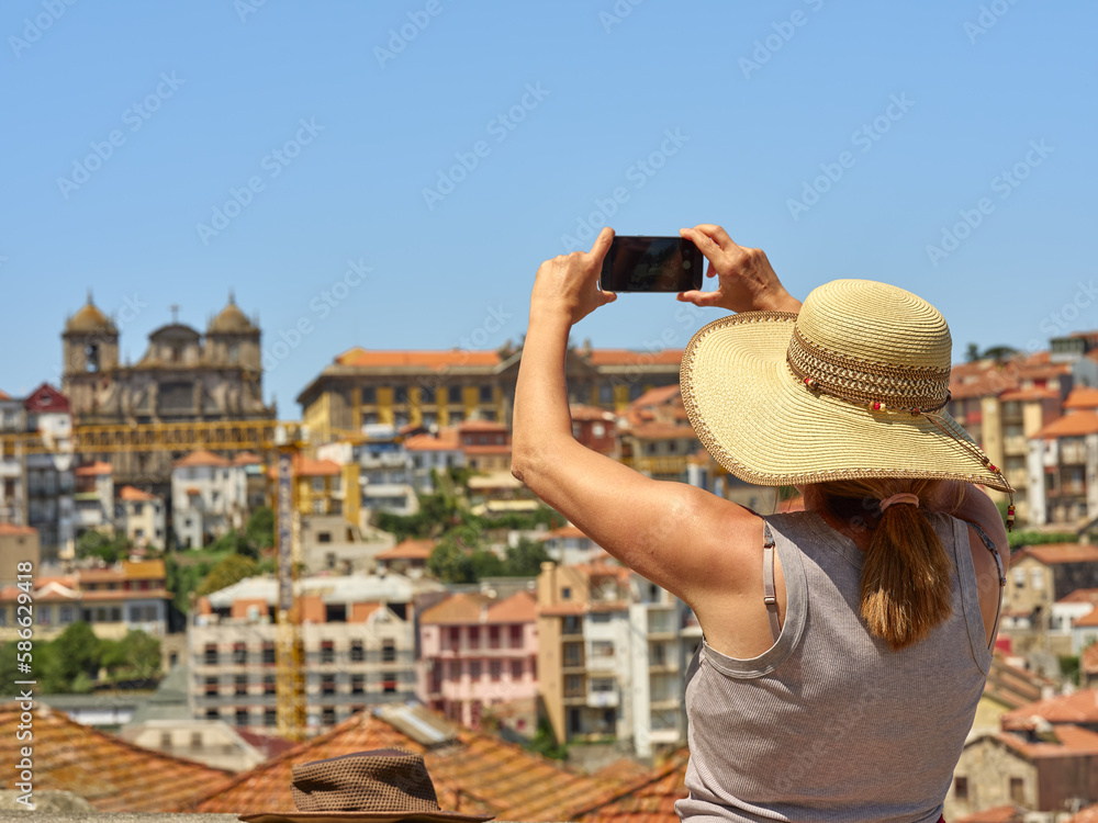 Woman wearing a straw hat to protect herself from the sun photographing the panorama of the historic city of Porto, Portugal. In the background the Torre de los Clérigos