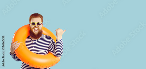 Photography Studio shot of funny ginger man in striped swimsuit, sun glasses and orange inflatable ring pointing at blank empty copy space background on right side