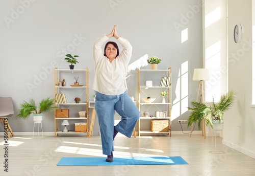 Relaxed plus size woman practicing yoga at home. Overweight woman standing on mat on one leg doing tree pose, keeping balance or meditating. Healthy lifestyle, wellness concept