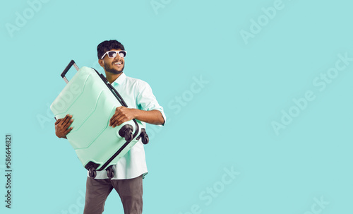 Happy excited indian guy tourist or vacationer holding travel suitcase on light blue background. Joyful ethnic young man looks towards copy space and smiles. Web banner. photo