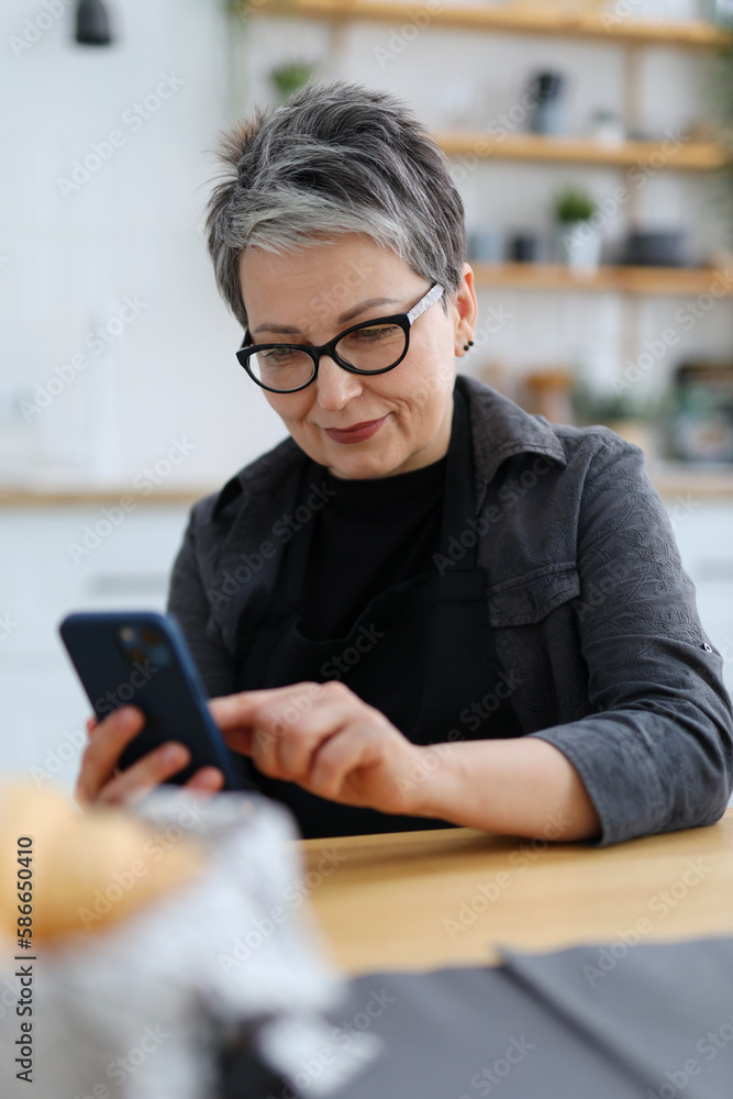 Mature woman in dark clothes looks at a smartphone while sitting at a table in the kitchen, close-up portrait. Mature woman in dark clothes looks at a smartphone while sitting at a table in the kitchen, close-up portrait.