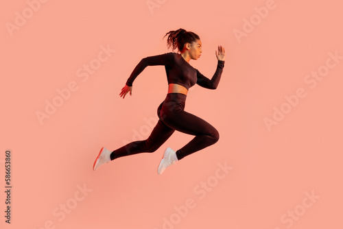 Sporty black lady jumping or running, posing in mid-air, exercising over peach neon background, side view, full length