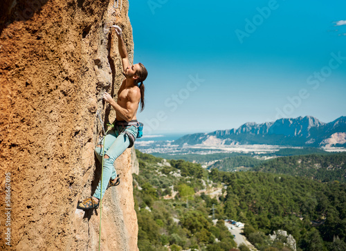 Strong muscular body man rock climber climbing on height vertical rock with rope on a mountains and blue sky background. Outdoor sports and recreation