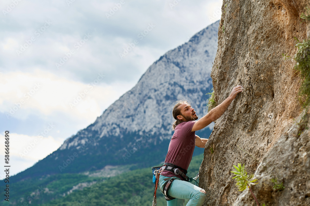 Active young man rock climber gripping hold, working on challenging route on cliff, beautiful mountains view on background