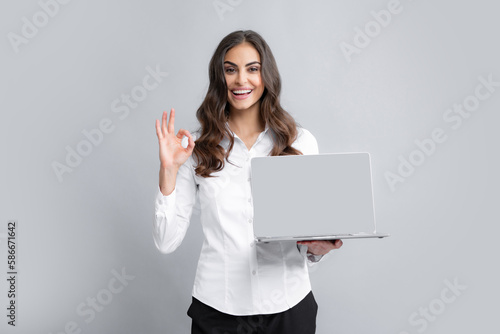 Cheerful business woman standing over grey wall with laptop computer. Portrait of pretty, charming, stylish, clever woman with open laptop chatting, using internet, isolated on grey background.