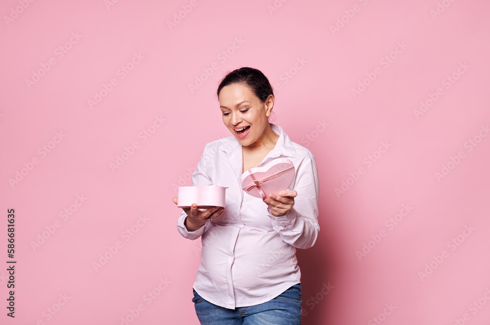 Smiling happy multi-ethnic pregnant woman, wearing casual white shirt and blue denim jeans, expressing happiness and positive emotion, rejoicing at present for Mother's Day on pink isolated background Smiling happy multi-ethnic pregnant woman, wearing casual white shirt and blue denim jeans, expressing happiness and positive emotion, rejoicing at present for Mother's Day on pink isolated background