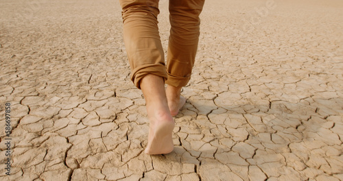 Close up shot of feet of adult man walking barefoot on bottom of dried lake or river, stepping on cracked soil ground destroyed by erosions - ecological issues concept 