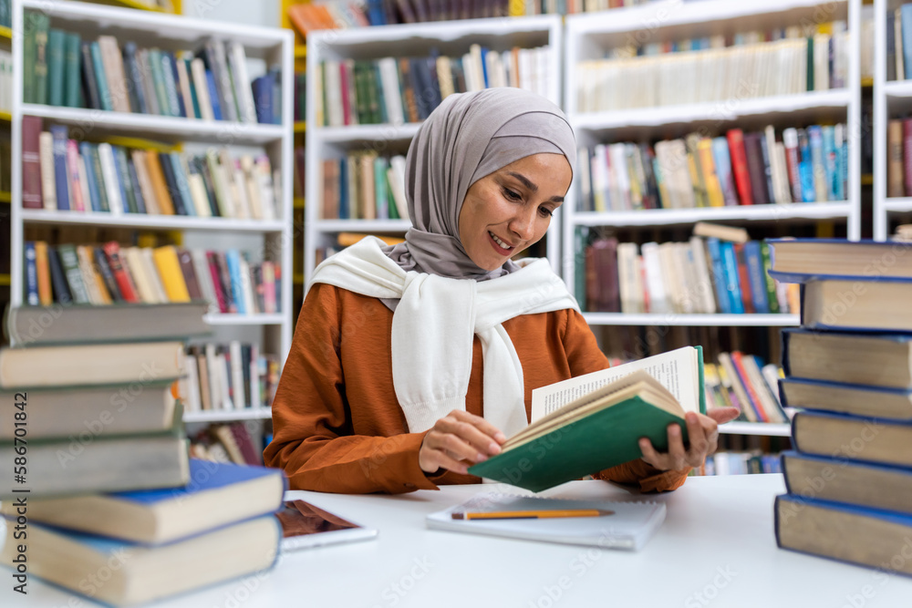 Young beautiful female student studying in the library, female student in hijab smiling among books studying material independently reading literature. Young beautiful female student studying in the library, female student in hijab smiling among books studying material independently reading literature.