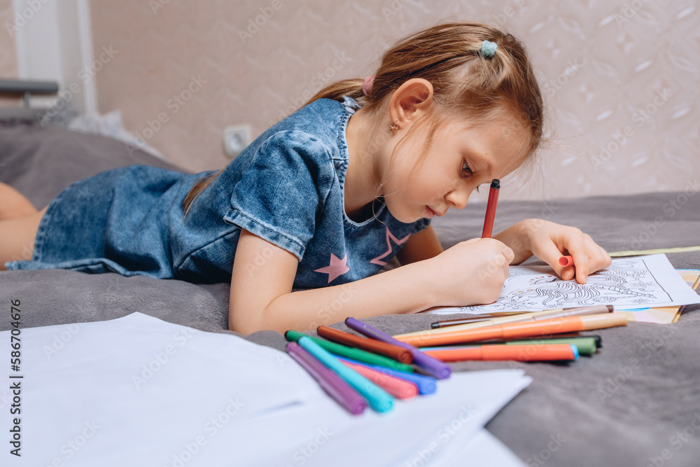 A little preschooler is lying on a soft bed and is engaged in coloring various pictures with felt-tip pens. A little preschooler is lying on a soft bed and is engaged in coloring various pictures with felt-tip pens.