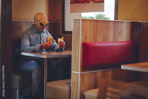 One man with alien mask eating alone inside a fast food store bad food hamburger and french fries. Ufo are among us living as human concept funny image. Invasion and diversity people indoor leisure photo