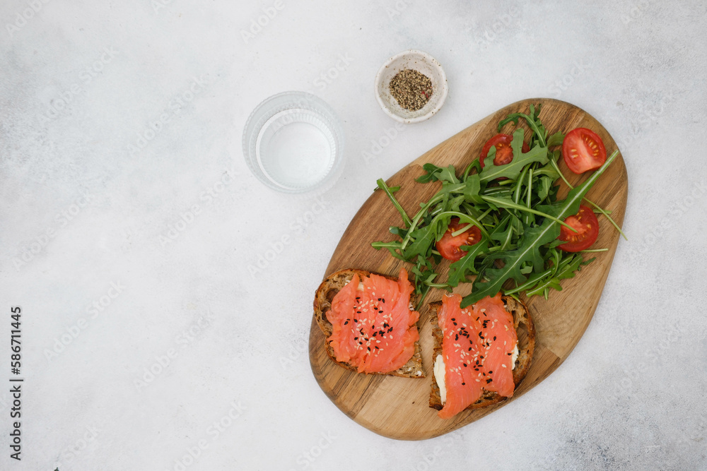 A wooden plate with salmon sandwiches, arugula, tomatoes, with glass of water on grey background. Healthy food. Horizontal frame.