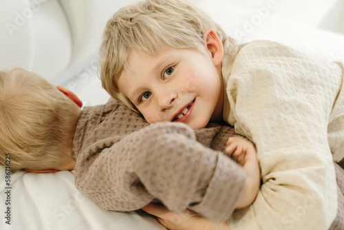 The brothers lie in dressing gowns in a white bed, play, hug, smile.