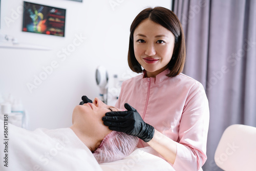 Cheerful Asian beauty specialist doing facial massage in clinic