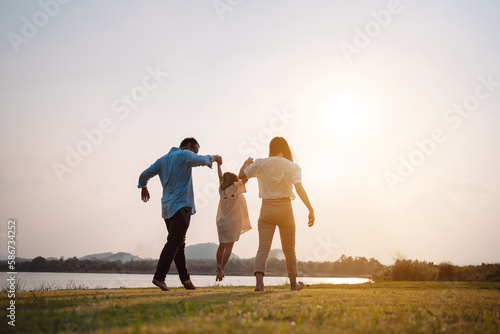 Fototapeta Naklejka Na Ścianę i Meble -  Happy family in the park sunset light. family on weekend running together in the meadow with river Parents hold the child hands.health life insurance plan concept.