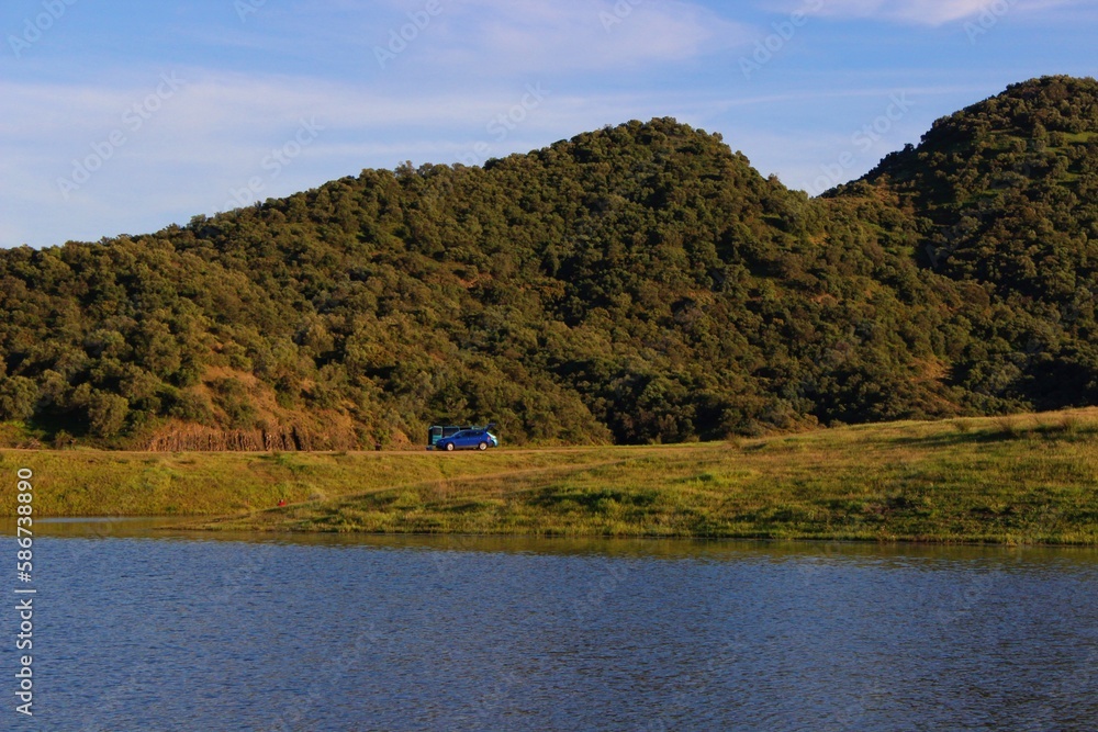 Landscape view of a lake and mountains in Cordoba, Spain. Camping area with two cars. Green, blue, yellow and brown colors. Reflection of the mountains in the water in summer