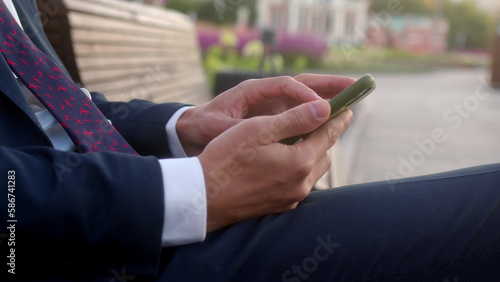 Close up young busy businessman sits on bench with purple flowers in flower beds behind him and use mobile phone. man in formal suit and tie is texting on smartphone  looking for information  trader