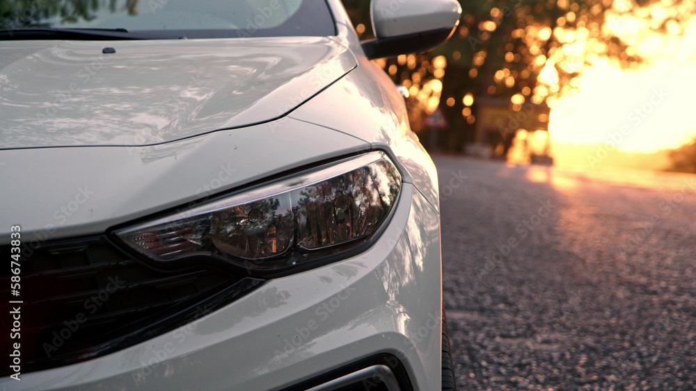 Close up view of front light of car headlights. unrecognizable white car is parked on side of road with setting sun in background. detail of exterior of modern car.