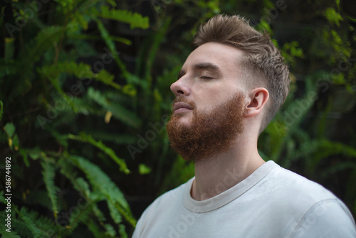 Portrait of happy young bearded man with beard breathing deep, inhale fresh air on natural background, green wall with leaves, flower in tropical country in garden. Eco, ecological concept. 