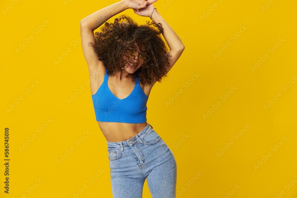 Woman with curly afro hair in a blue T-shirt on a yellow background dancing flying hair with sunglasses yellow, hand signs, look into the camera, smile with teeth and happiness, copy space Woman with curly afro hair in a blue T-shirt on a yellow background dancing flying hair with sunglasses yellow, hand signs, look into the camera, smile with teeth and happiness, copy space