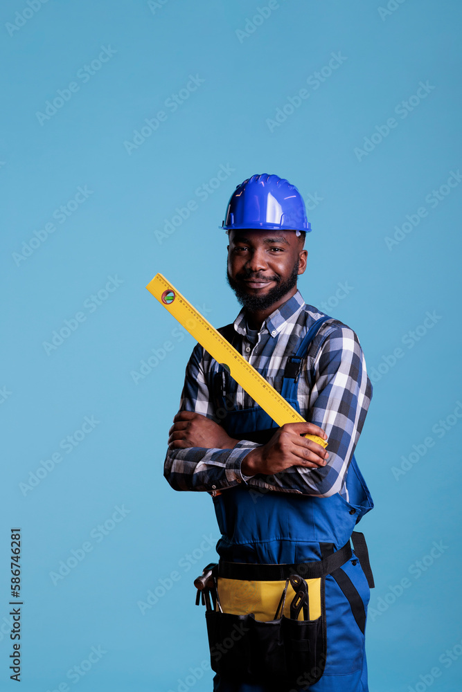 Smiling african american repair worker in uniform holding tape measure on blue studio background. Experienced builder holding work tools wearing coveralls and hard hat. Smiling african american repair worker in uniform holding tape measure on blue studio background. Experienced builder holding work tools wearing coveralls and hard hat.