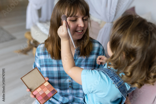 Pretty cute little girl doing make up for her mother with eye shadows palette, brush. Quality family time, enjoying domestic life, motherhood. Women's or Mother's Day celebration, having fun together