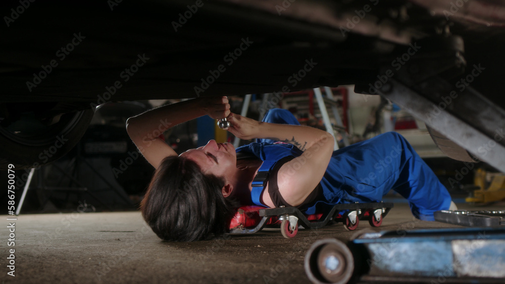woman mechanic with tattoos lies under car on car repair trolley and repairs it with tools. View of girl in blue overalls in car repair area, who performs vehicle maintenance in car service.