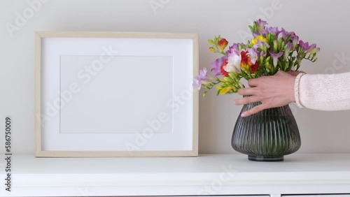 Female hands puts flowers in a vase. Wooden frame mockup in white room interior, empty mock-up with copy space. Light frame with a vase in the interior for design presentation.