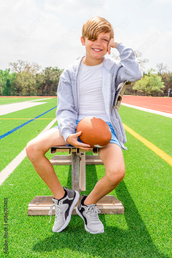 Smiling preteen boy with football sitting relaxing on bench at a sports ...