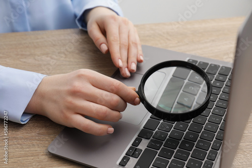 Woman holding magnifier near laptop at wooden table, closeup. Online searching concept