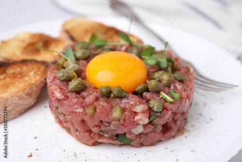 Tasty beef steak tartare served with yolk, capers, toasted bread and greens on plate, closeup