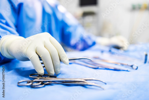 Hand of doctor or surgeon in blue gown pick up surgical clamp instrument or tools inside operating theatre with blur background.Medical equipment for surgery inside room in hospital.Modern technology.