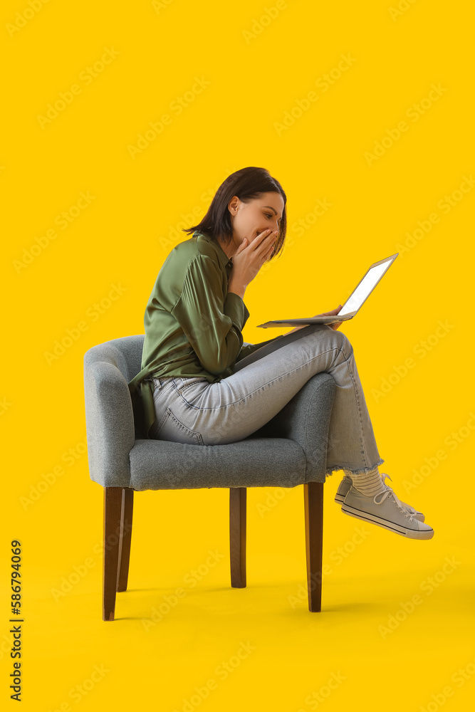 Beautiful woman using laptop in soft armchair on yellow background