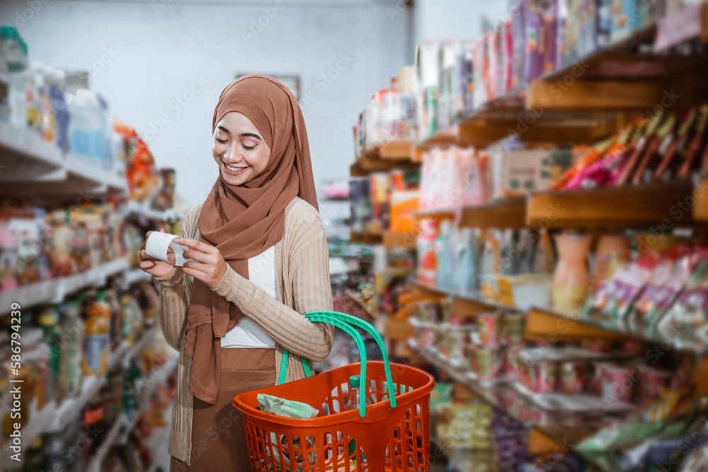 Muslim woman checking shopping receipt while carrying basket in supermarket