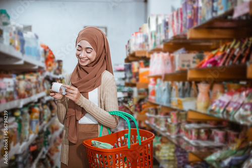 Muslim woman checking shopping receipt while carrying basket in supermarket