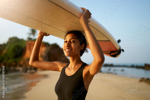 African american woman standing with surfboard on head on ocean beach. Black female surfer posing with surf board. Pretty multiethnic girl goes on surfing session on tropical location.