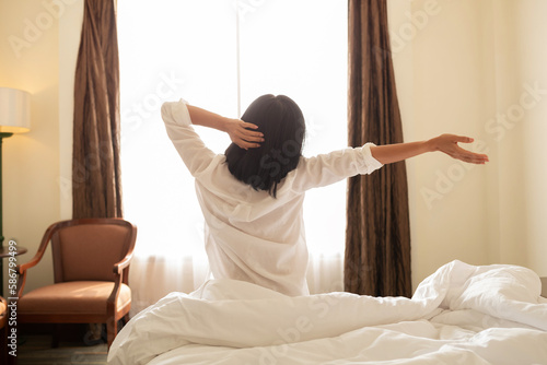 Asian woman wake up in the morning, sitting on white bed and stretching, feeling happy and fresh. happy woman lying on soft pillows awaken in cozy bedroom, relaxation concept.