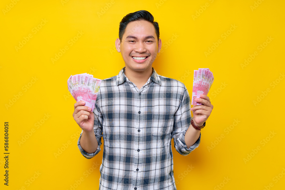 Portrait of cheerful young Asian man wearing plaid shirt Holding fan of cash money in rupiah banknotes isolated on yellow background. people lifestyle concept Portrait of cheerful young Asian man wearing plaid shirt Holding fan of cash money in rupiah banknotes isolated on yellow background. people lifestyle concept