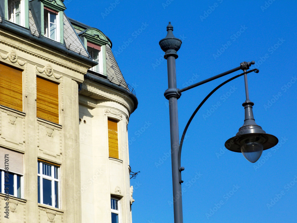 retro style lamppost head. closeup detail. low angle view. stucco building facade beyond. glass lens. gray lamp hood. residential neighborhood. mansard roof. yellow wood roll down window shutters retro style lamppost head. closeup detail. low angle view. stucco building facade beyond. glass lens. gray lamp hood. residential neighborhood. mansard roof. yellow wood roll down window shutters