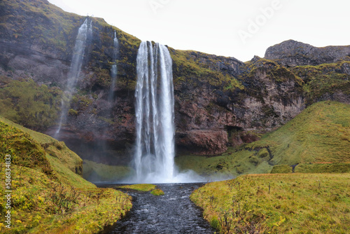 A majestic waterfall in Iceland cascades down a mossy cliff into a tranquil stream below, surrounded by lush green hills, showcasing the natural beauty of the Icelandic landscape