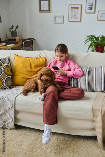 Vertical image of little girl sitting on sofa in the room and taking photo of her dog on smartphone